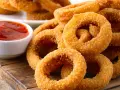 Aros de cebolla dorados y crujientes apilados junto a un tazón de salsa de tomate.