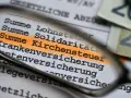 Lupe über Dokument mit hervorgehobener Kirchensteuer. Geld liegt daneben.