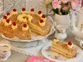 Ein köstlicher Frankfurter Kranz, verziert mit Buttercreme-Tupfern und Kirschen, umgeben von gehackten Nüssen. Ein Stück ist auf einem Teller angerichtet.