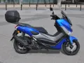 Niebieski skuter Yamaha NMAX 125 z kufrem, gotowy do jazdy. Jego prędkość maksymalna pozwoli na szybkie przemieszczanie się po mieście.
