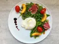 Burrata mit Rucola, Tomaten, Pfirsichspalten und Prosciutto auf einem Teller.