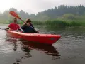 Para płynie kajakiem po rzece, w tle las i trzciny. To piękny dzień na spływ Pilicą, miasto w oddali.