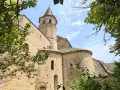 Vue d'une église romane à Villeneuve de Rouergue, encadrée par des arbres verdoyants sous un ciel bleu parsemé de nuages.