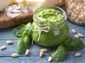 Słoik domowego pesto z bazylii, otoczony świeżymi liśćmi bazylii i orzeszkami pinii. Obok kawałki parmezanu. Idealne do makaronu.