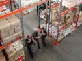 Magazzino con scaffalature piene di scatoloni e taniche. Tre persone camminano velocemente, forse per svolgere mansioni di aiuto magazziniere.