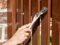 Ręka maluje drewniany płot pędzlem. Decyzja: podbitka lakierobejca czy impregnat?