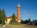 Wysoka, ceglana latarnia morska Jarosławiec góruje nad placem z drzewami i budynkiem.