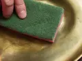 Mano che pulisce un piatto d'ottone con una spugna verde. Scopri come pulire l'ottone fai da te per farlo brillare.
