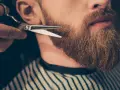 Un barbero recorta una barba larga y rojiza con tijeras.