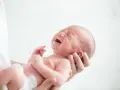 Noworodek z zamkniętymi oczami i otwartymi ustami, wyraźnie cierpiący z powodu kolki jelitowej, trzymany w ramionach.