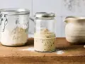 Blubbernder Sauerteig im Glas, bereit für schnelles Sauerteig herstellen. Daneben ein Glas mit Mehl und ein Krug.