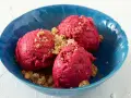 Drei Kugeln leuchtend pinkes Sauerrahm Eis, bestreut mit knusprigen Krümeln, in einer blauen Schale.