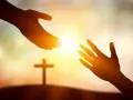 Zwei Hände reichen sich im warmen Licht entgegen, ein Kreuz im Hintergrund symbolisiert die Hilfe von Jesus Christus.