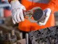 Mechanik w białych rękawicach trzyma tłok z widocznymi objawami zużytych pierścieni tłokowych. Obok leży głowica silnika.