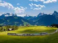 A train travels through a lush green valley with snow-capped mountains in the background. Many positive goldenpass line reviews mention this scenic route.