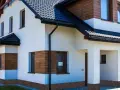 Nowoczesny dom z białym tynkiem i drewnianymi akcentami. Z czego zrobić cokół wokół domu? Widać tu kamyczki, które mogą być inspiracją.