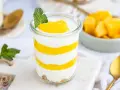 Schichtweise geschichtetes Mango-Parfait in einem Glas, garniert mit Minze. Daneben eine Schüssel mit frischen Mangostücken.