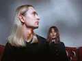 Łukasz Rozmysłowski z długimi blond włosami i kobieta z brązowymi włosami siedzą na skórzanej kanapie.