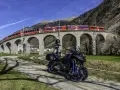 Motociclista e moto su strada sterrata, con un treno rosso che attraversa un viadotto ad archi in pietra. Paesaggio alpino con montagne sullo sfondo, vicino al passo della Novena.