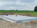 Gotowa płyta fundamentowa na budowie, otoczona piaskiem i zielenią. W tle widać pola i budynki przemysłowe.