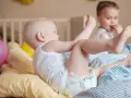 Dzieci bawią się na łóżku. Maluch w pieluszce sięga po zabawkę, a starszy chłopiec je. Ciekawe, ile pieluch miesięcznie zużywają takie energiczne maluchy!