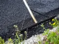 Verktyg sprider ny asfalt för att asfaltera uppfart. Grönska och gula blommor växer vid sidan.