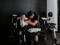 Man in black shirt stretches his outer hamstring by holding his foot, surrounded by stationary bikes in a gym.