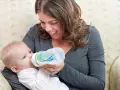 Uśmiechnięta mama karmi niemowlę butelką z systemem odpowietrzania. Smoczek do butelki Dr Brown zapewnia komfortowe karmienie.