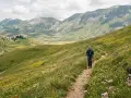 Escursionista su sentiero fiorito nel **Val di Norcia**, con borgo in lontananza e montagne verdi.