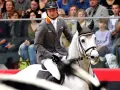 Un cavalier en tenue grise monte un cheval blanc, prêt pour la compétition. Le public est attentif.