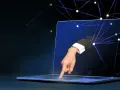 Ręka w garniturze wychodzi z ekranu laptopa, dotykając klawiatury. Sieć połączeń symbolizuje zdalny dostęp do komputera.