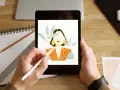 Ręce trzymają tablet z rysunkiem kobiety, tworzonym w aplikacji procreate. Obok leżą szkicownik i ołówek.