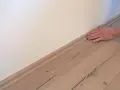 Hand drückt Sockelleisten an die Wand. Der Boden ist aus hellem Holz.