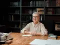 Uśmiechnięta starsza pani w okularach siedzi przy biurku, na którym leży notatnik i maszyna do pisania. Za nią regały pełne książek, jakby przygotowywała się do obrony swojego świadectwa dojrzałości.