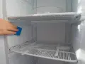 A hand uses a blue sponge to clean ice buildup inside a fridge freezer, showing a method for how to defrost fridge freezer without turning it off.