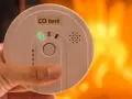 Detektor czadu z zieloną diodą i napisem "CO test" na tle płomieni.