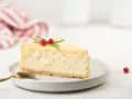 Una fetta di cheesecake con yogurt greco, guarnita con rosmarino e bacche rosse, su un piatto bianco con cucchiaino dorato.