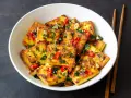 Chrupiące kawałki tofu satay, posypane chili, sezamem i ziołami, podane w białej misce z pałeczkami.