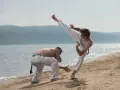 Dwóch mężczyzn ćwiczy capoeirę na plaży. Jeden wykonuje wysokie kopnięcie, drugi jest w pozycji obronnej. Widać ich płynny ginga styl gry.