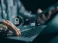 Manos sobre teclado de laptop, con iconos de seguridad digital: candado, huella, base de datos, globo terráqueo. Esto ilustra qué es la seguridad informática.