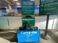 Frontier Airlines personal item size guide at the airport, showing a tote bag fitting in the "Personal Item" box and a roller bag in the "Carry-On" box.