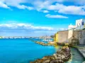 Un incantevole cammino del Salento: un porto con barche a vela, case bianche su scogliere dorate e un mare azzurro sotto un cielo limpido.