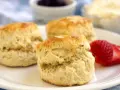 Tre gyllene engelska scones på en vit tallrik med en jordgubbe, sylt och grädde i bakgrunden.
