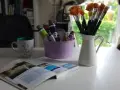 Pędzelki malarskie w dzbanku, tubki farb w fioletowym wiaderku, kubek z napisem i otwarta książka.