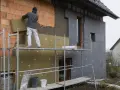 Pracownik montuje izolację na elewacji domu. Widać ceglaną ścianę, wełnę mineralną i szare płyty izolacyjne.