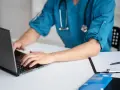 Lekarz medycyny pracy na laptopie, przygotowuje dokumentację. Zastanawiasz się, ile trwa wizyta u lekarza medycyny pracy?