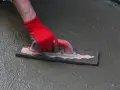 Ręka w czerwonej rękawicy wygładza cienką wylewkę na starym betonie za pomocą kielni.