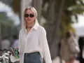 Donna con occhiali da sole, collana, camicia bianca e jeans, tiene in mano una lattina verde e un telefono.