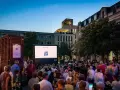 Ein lauer Sommerabend in Berlin: Menschen genießen ein Open-Air-Kino-Erlebnis vor einer Leinwand mit dem Logo "Lux Augenoptik".