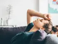 Eine Frau liegt erschöpft auf einem Sofa, die Hand auf der Stirn. Sie verarbeitet den tod der schwiegermutter.